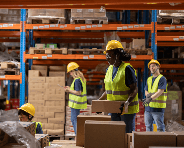 Team of warehouse workers wearing safety gear, including hard hats and vests, efficiently organizing and transporting boxes in a large warehouse, showcasing teamwork in warehouse operations and inventory management.
